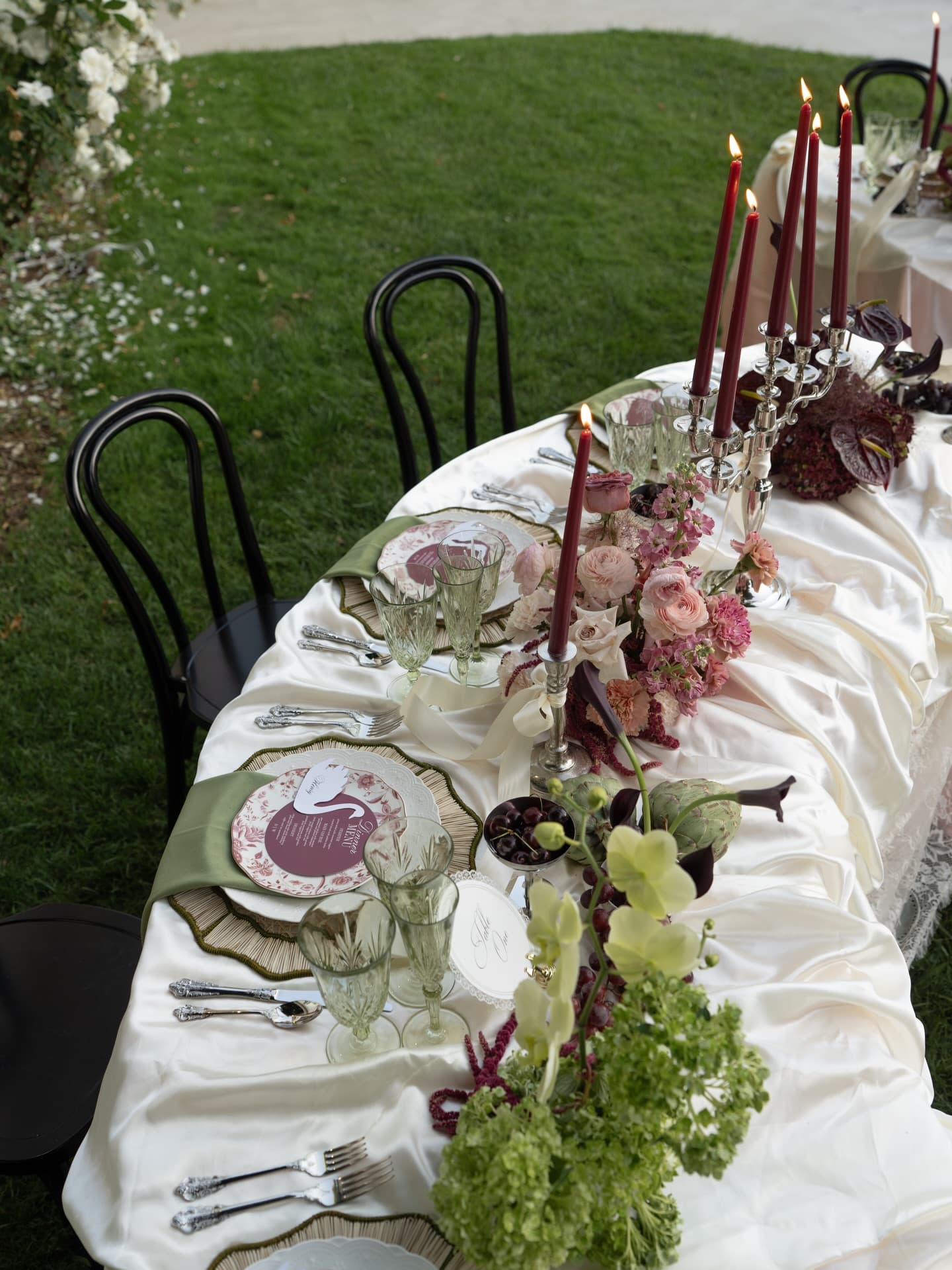 An olive velvet runner and gold-rimmed charger on champagne satin, with green hydrangea and a pink dinner menu, seen close