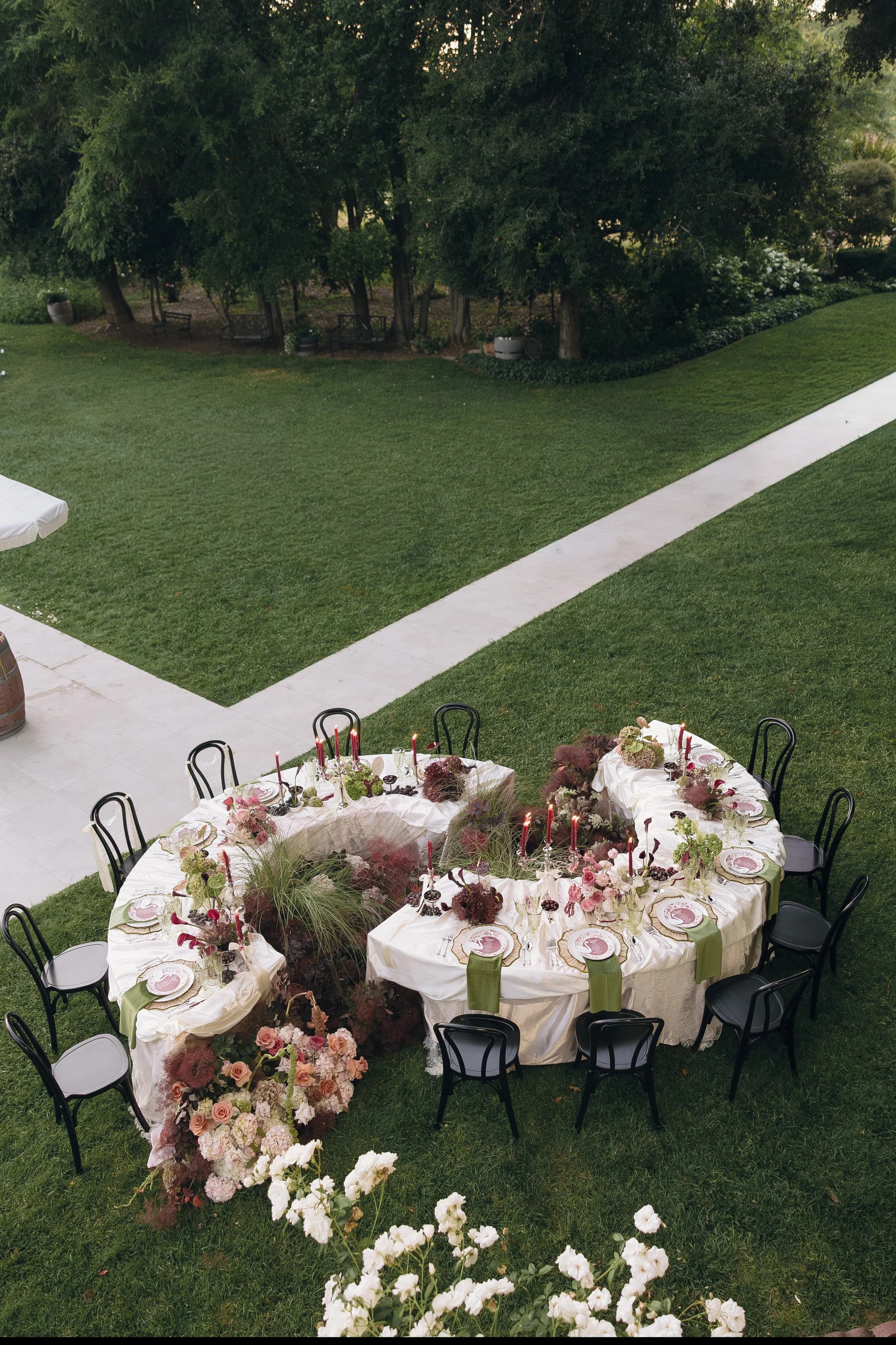 A serpentine dinner table on a lawn seen from above, set with florals at both ends and ringed with black bentwood chairs, garden paths beyond